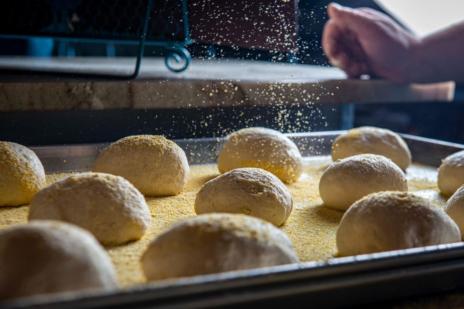 Dough balls on a tray being dusted with a falling shower of coarse flour
