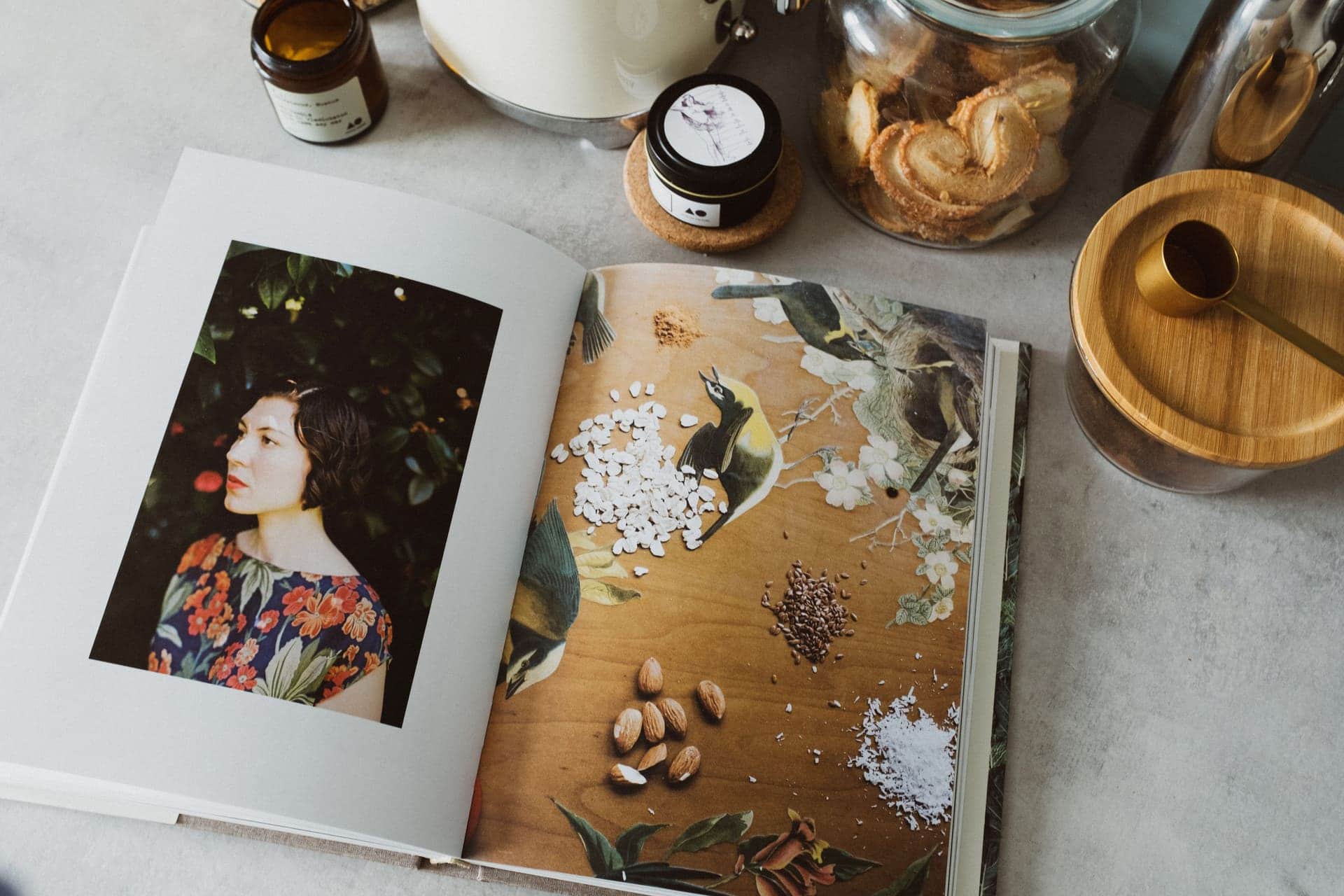 An open cookbook lying on a pale kitchen counter beside glass storage jars and a wooden lid