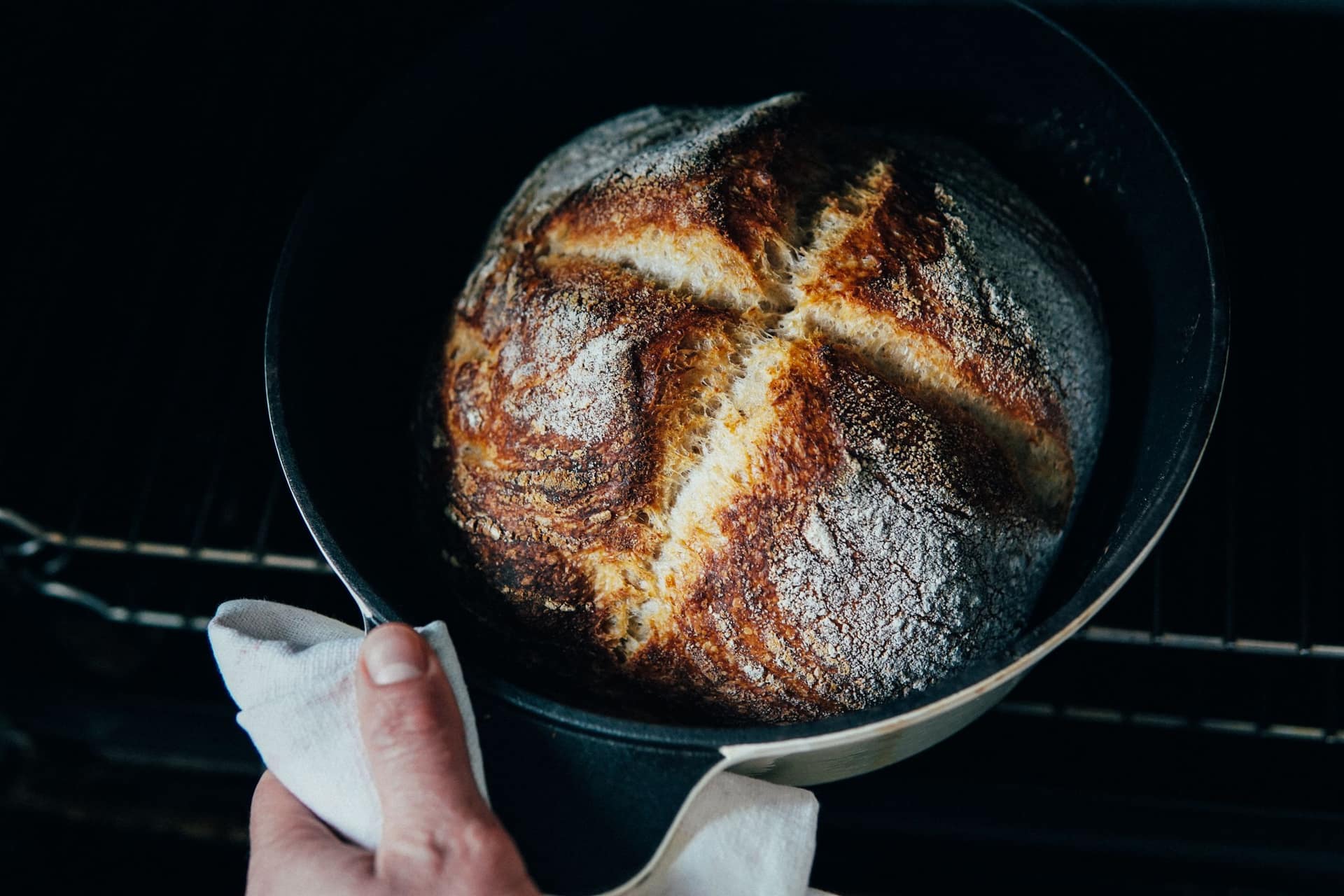A scored round loaf in a dark cast iron pot being lifted from the oven with a cloth