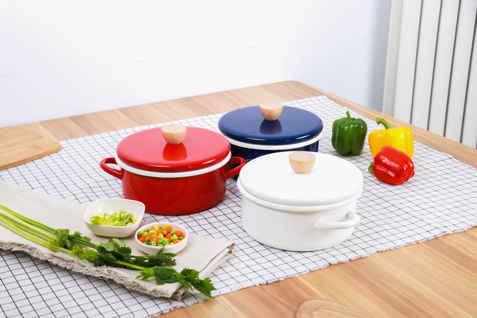 Three enameled pots with lids on a table beside fresh vegetables