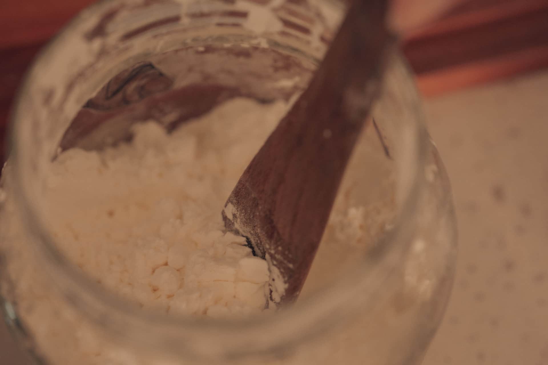 A wooden spoon stirring a pale mixture in a glass jar on a kitchen counter
