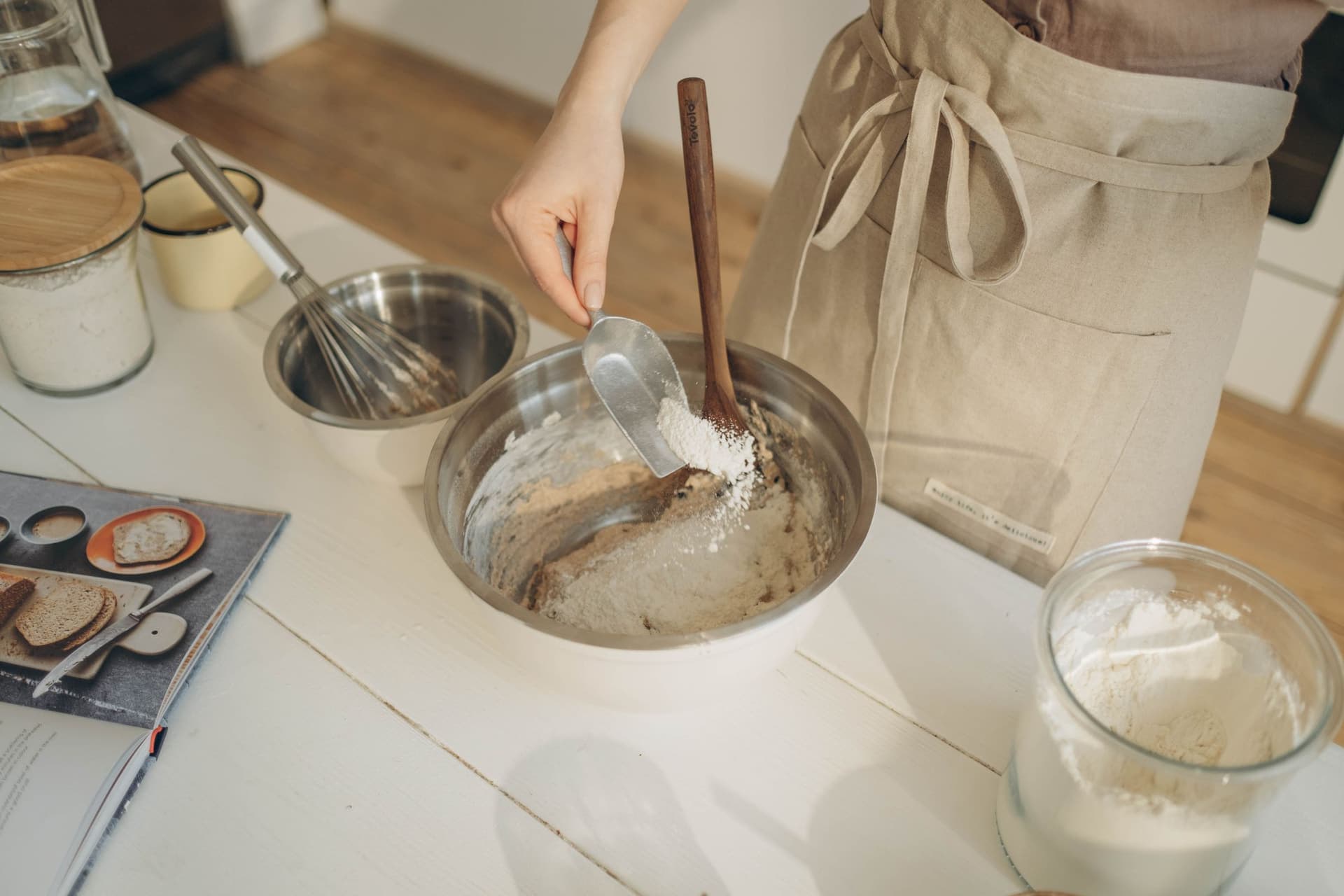 Flour being scooped into a metal mixing bowl of dough with a long wooden-handled mixer standing in it