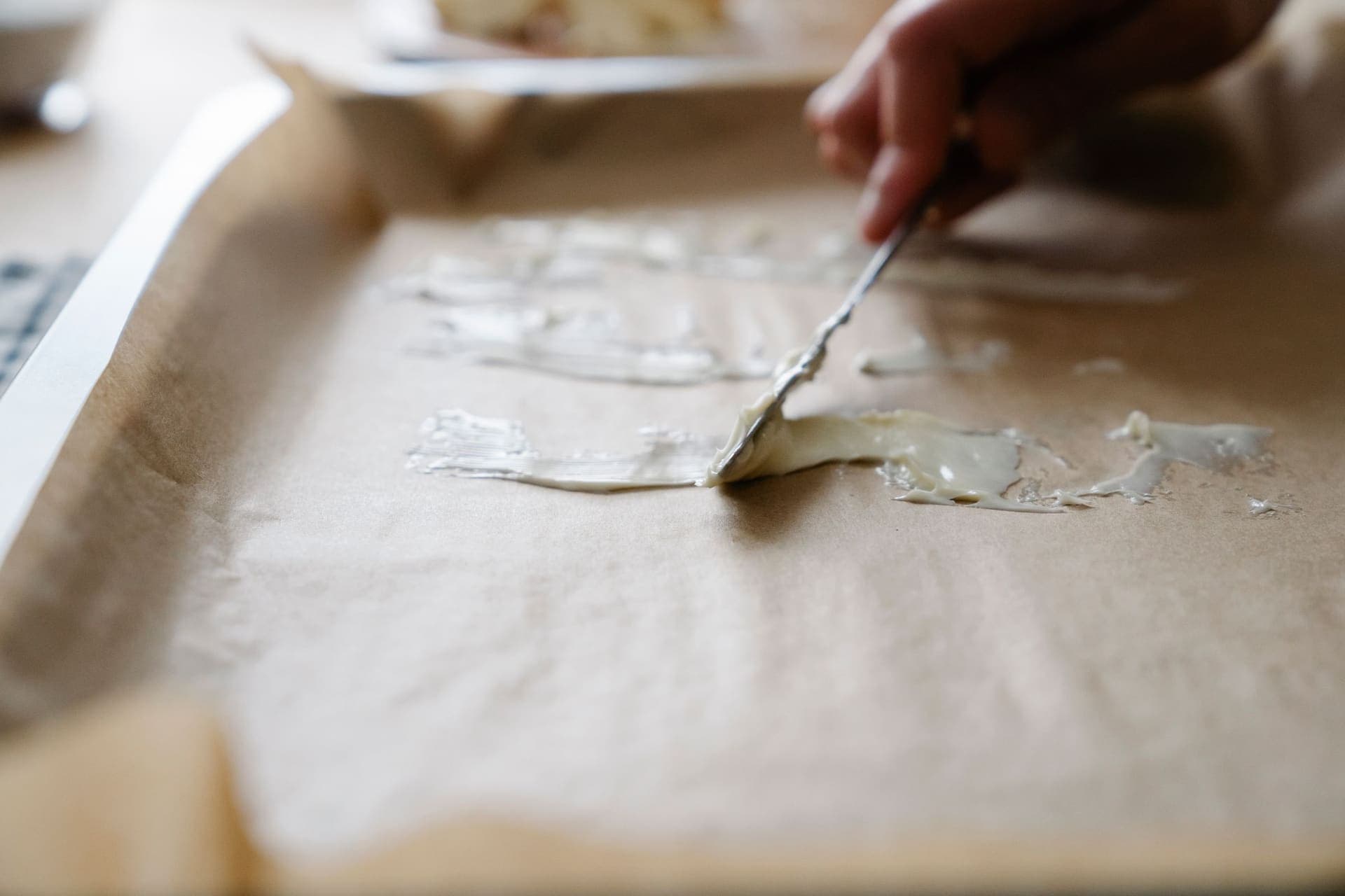 A hand spreading a thin layer of pale paste across a parchment-lined baking tray with a metal spatula