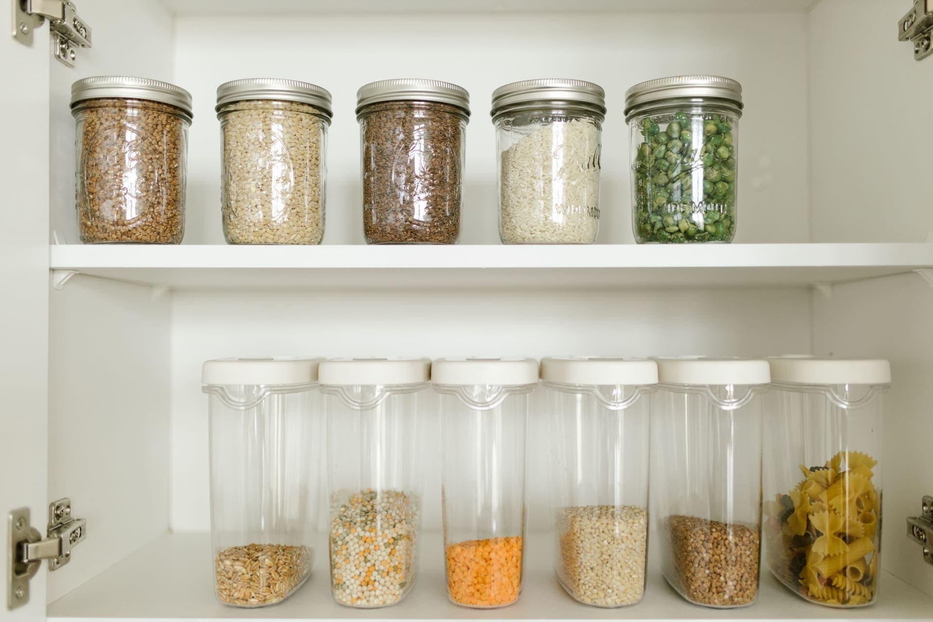 Glass jars and clear plastic containers of grains and pasta on white cupboard shelves