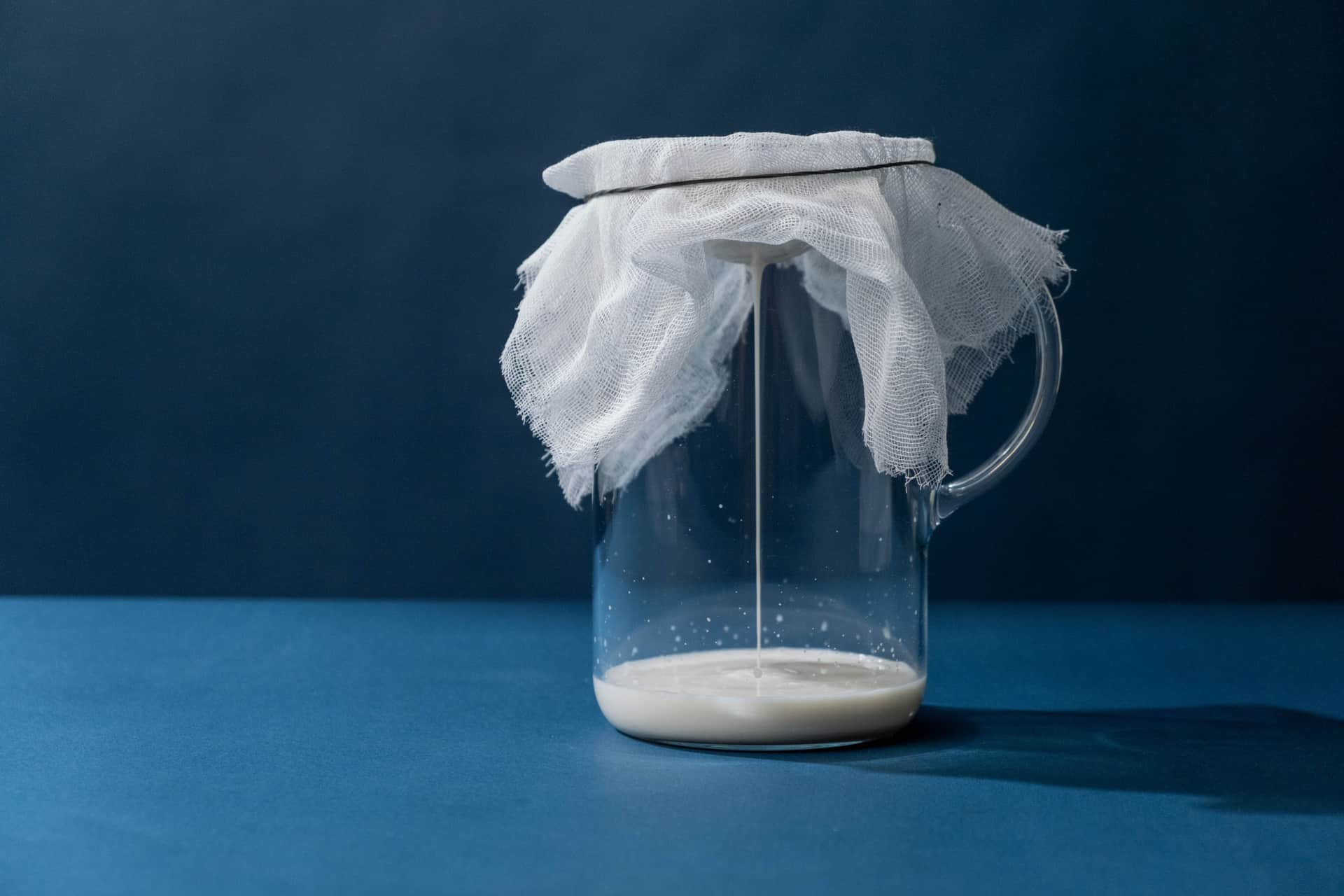 A glass pitcher covered with cheesecloth straining cultured milk