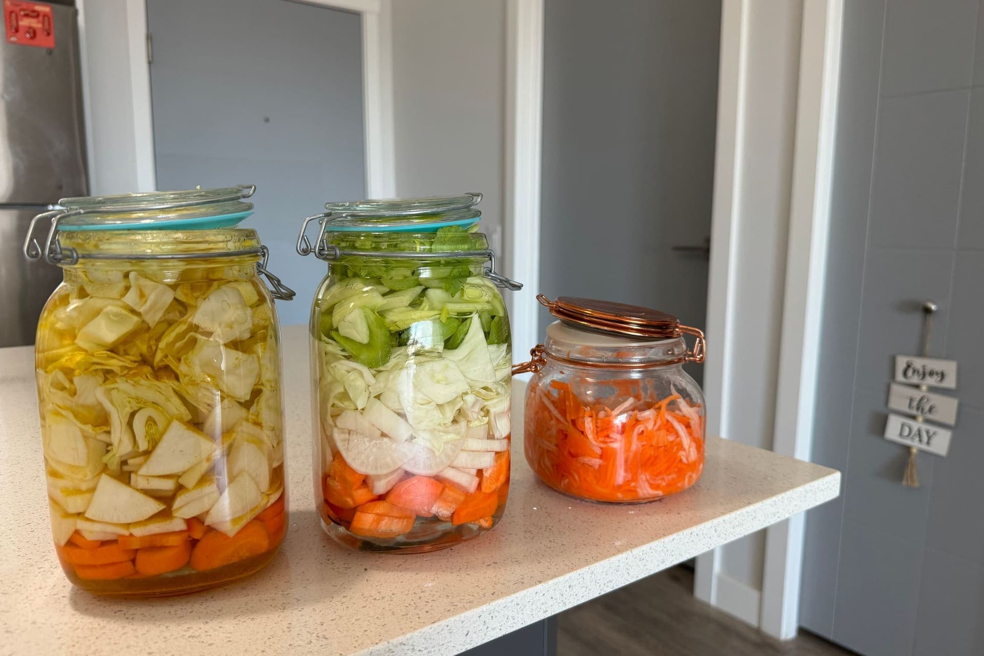 Three clamp-lid glass jars of cabbage, radish and carrot fermenting in brine on a kitchen counter