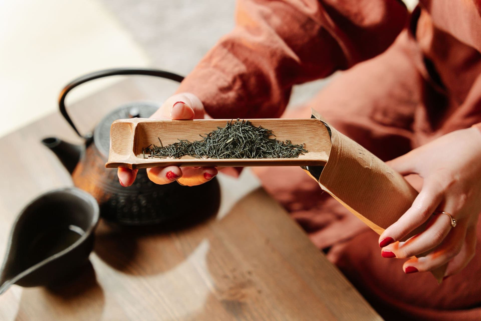 Hands holding a bamboo scoop of loose green tea leaves beside a cast iron teapot