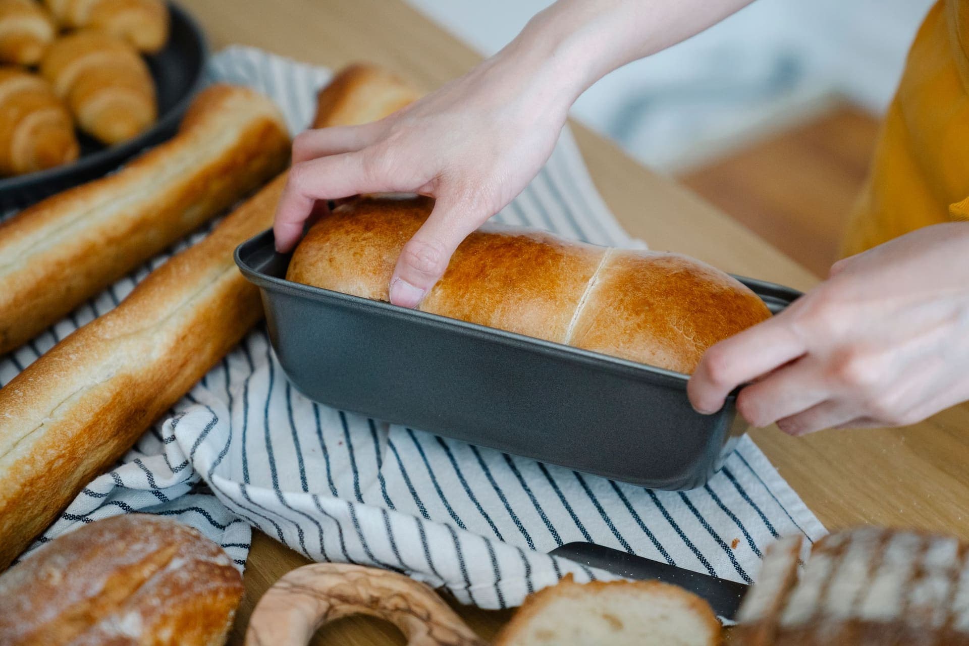 Hands holding a dark metal loaf pan with a golden domed sandwich loaf still in it