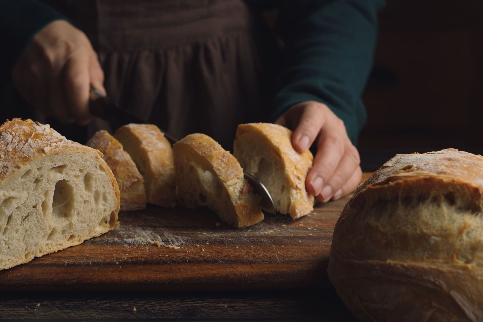 Hands slicing a round rustic loaf into even slices on a wooden board with a serrated knife